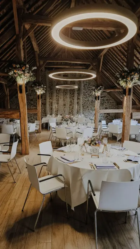 Décoration de mariage dans une grange : tables rondes, suspension lumineuse et compositions florales sur colonnes – Château de Florzée, mariage de G et C, septembre 2025