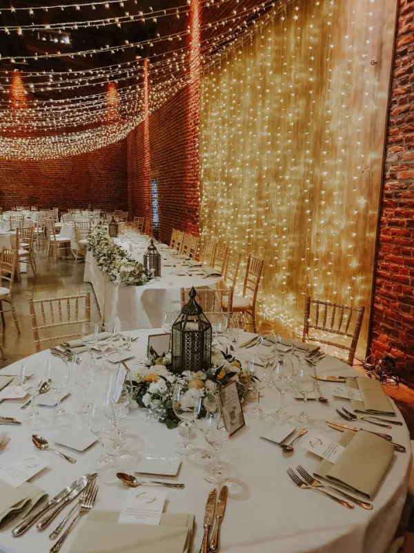 Salle de mariage en briques : rideau lumineux, lanternes et tables rondes – Ferme de Bertinchamps, mariage de S et G, juin 2025