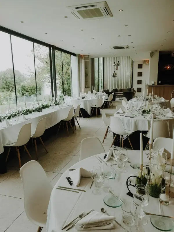 Salle lumineuse : longues tables, chaises blanches et chemins de feuillage – Kattebroek, mariage de F et G, août 2024