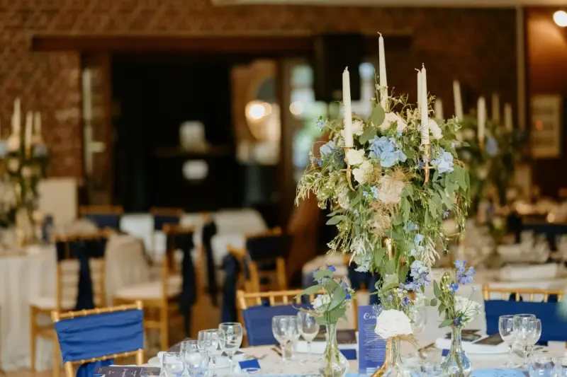 Centre de table sur chandelier : fleurs bleues et blanches, nœuds de chaise bleus – Ferme des Oliviers, mariage de M et M, juillet 2024