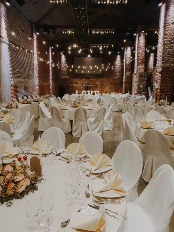 Grange en briques avec guirlandes lumineuses et housses de chaise blanches – Ferme de Bertinchamps, mariage de J et M, juillet 2024
