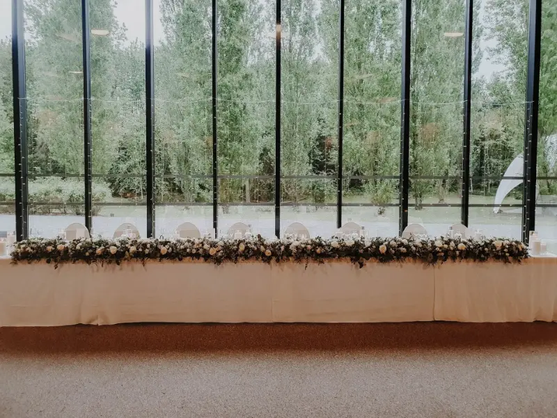 Table d'honneur devant des baies vitrées avec guirlande de feuillage – Jardins du Croisé Laroche, mariage de J et L, avril 2024