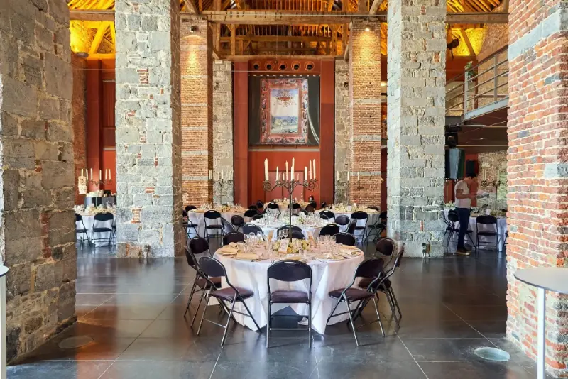 Salle en pierre et briques : tables rondes, chandeliers et chaises noires – Ferme de l'Abbaye de Moulins, mariage de M et H, avril 2024