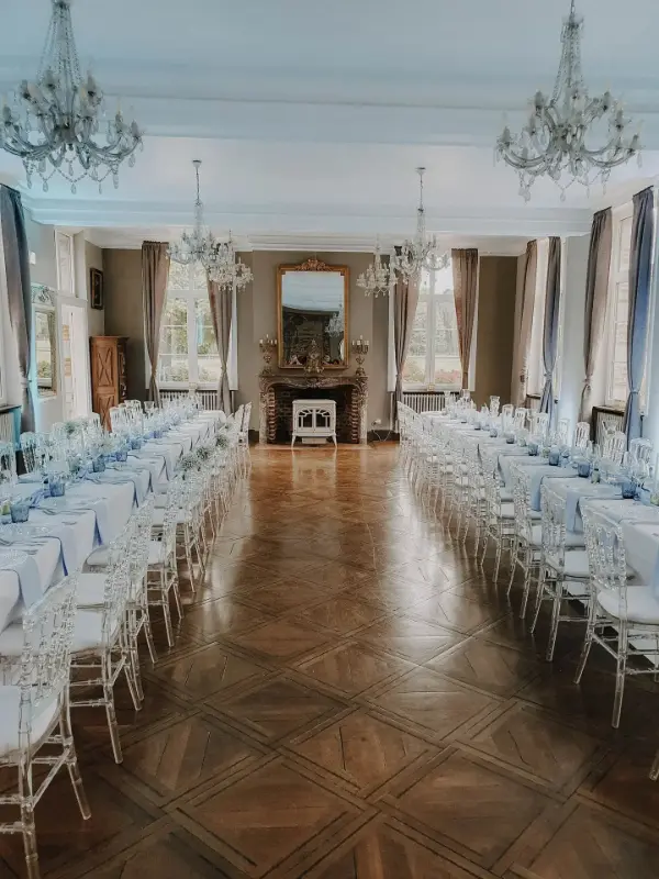 Salon de château avec cheminée et lustres : longues tables et chaises Napoléon blanches – Château de Thieusies, mariage de M et M, juillet 2023