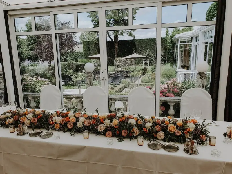 Table d'honneur avec chemin floral orange et pêche et housses de chaise blanches – Salons Leieburcht, mariage de C et G, juillet 2023