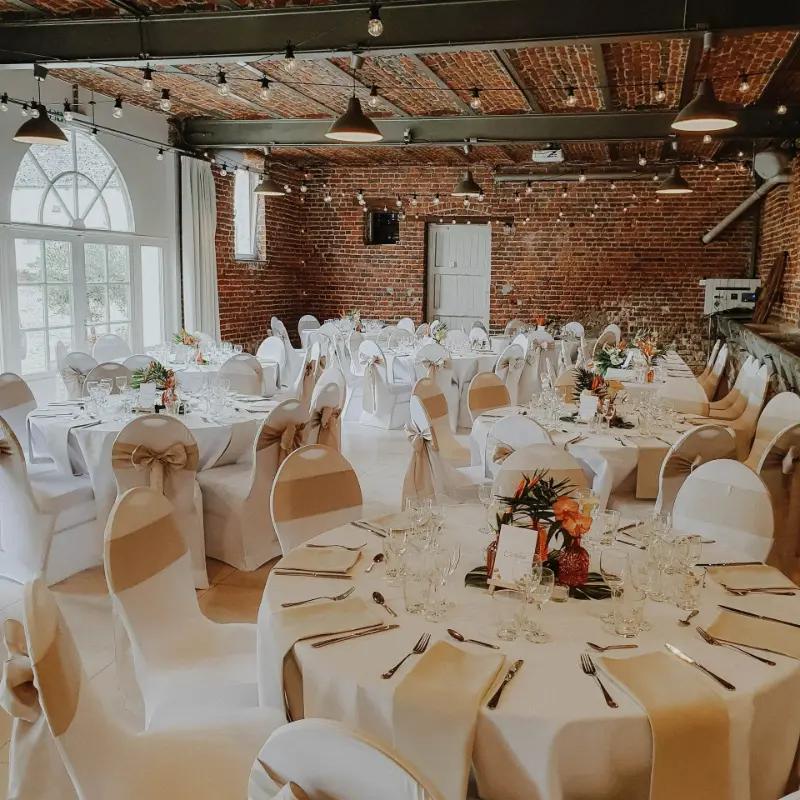 Salle en briques : housses de chaise blanches et nœuds dorés – Ferme de Bertinchamps, mariage de M et A, juin 2023