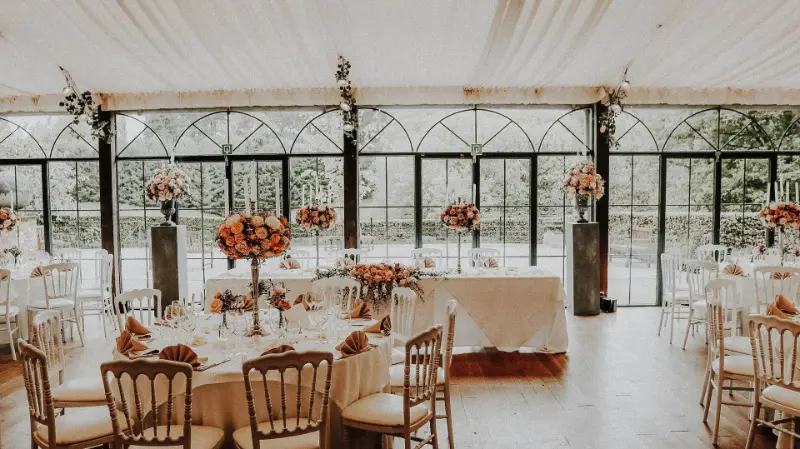 Véranda : tables rondes et compositions de fleurs orange sur hauts supports – Orangerie de Beauval, mariage de A et P, mai 2023
