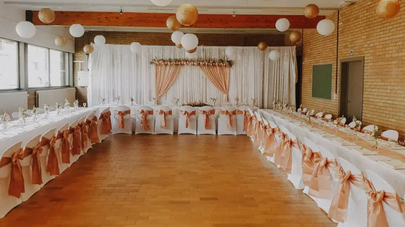 Tables en U avec nœuds de chaise saumon, voilages et lanternes en papier – Sporthal De Singel, mariage de P et G, avril 2022